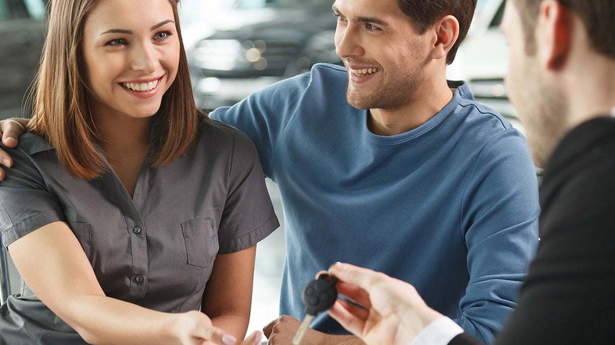 A smiling couple receives car keys from a salesperson. The woman looks at the keys, the man looks at the salesperson.