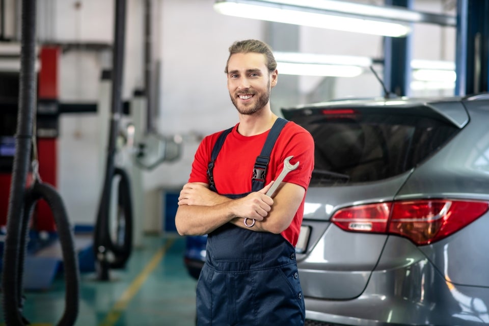 A smiling mechanic in a red shirt and blue overalls holding a wrench