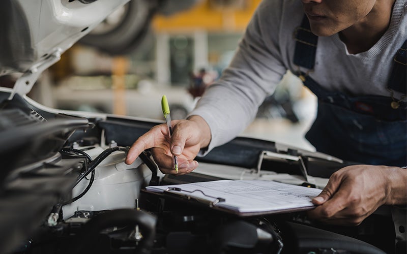 A mechanic holding a clipboard points at a car engine with a pen