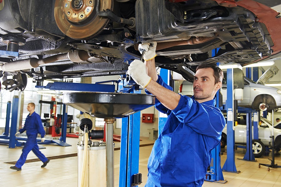 A mechanic in blue overalls and white gloves working under a car lifted on a hydraulic jack.